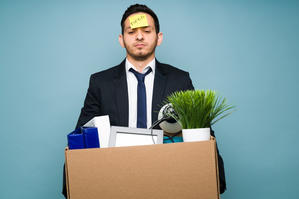 Sad and upset businessman in a suit holding a box with office supplies. The Hispanic man with a sticker shot on his forehead just lost his job